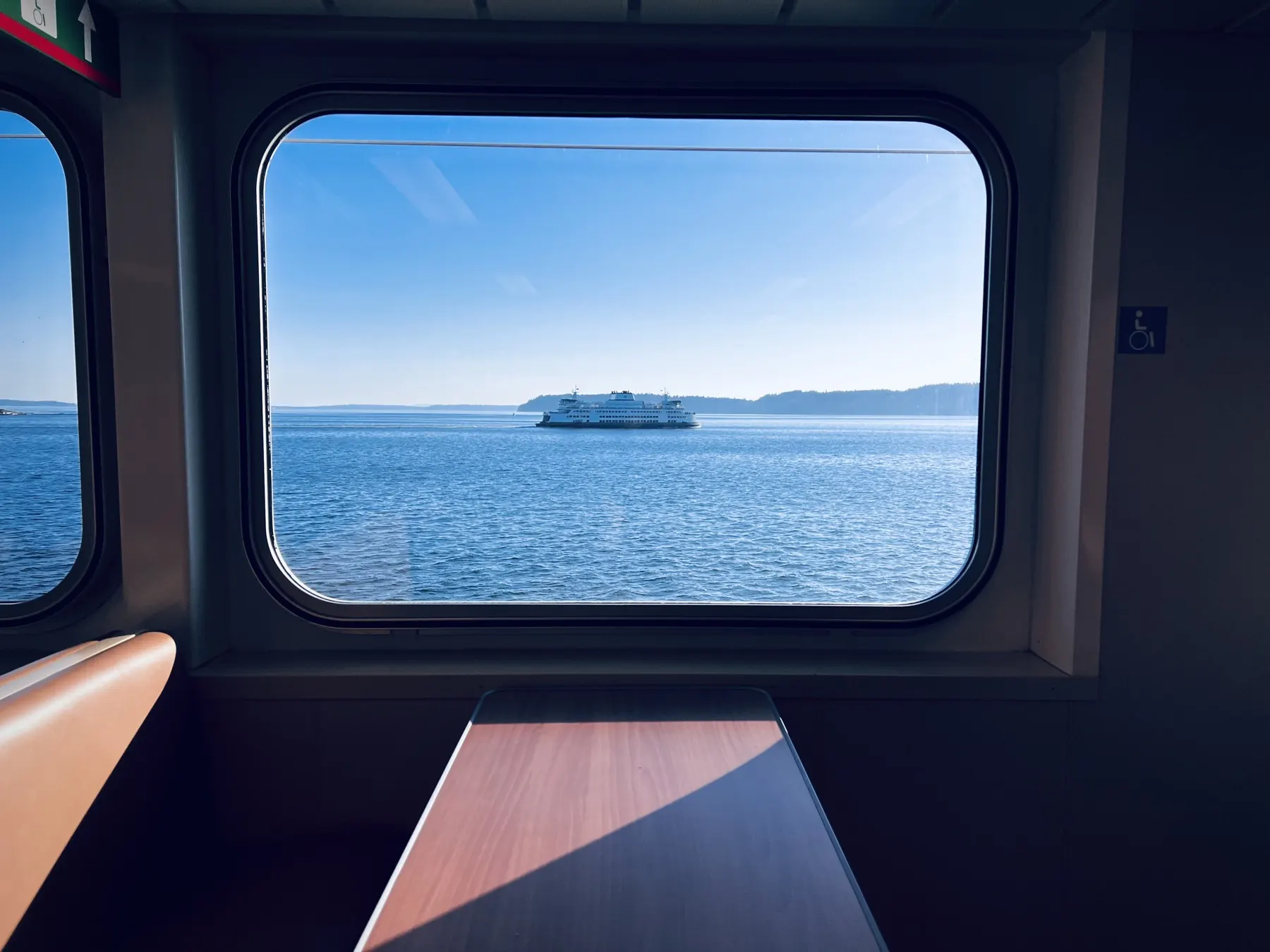 From inside a Washington State ferry: a big rounded window over Puget Sound, another ferry crossing in the distance, a table in the sun below.
